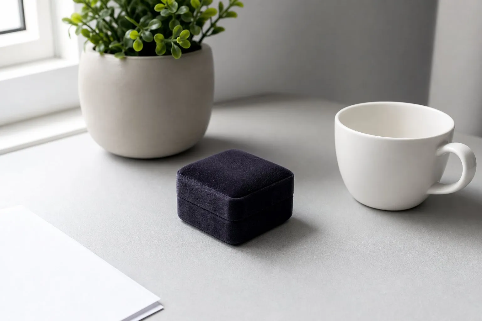A closed velvet ring box on a pale desk beside a plain ceramic cup