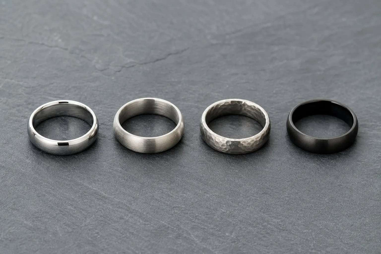 Four plain men's wedding bands in a row on slate, each with a different surface finish
