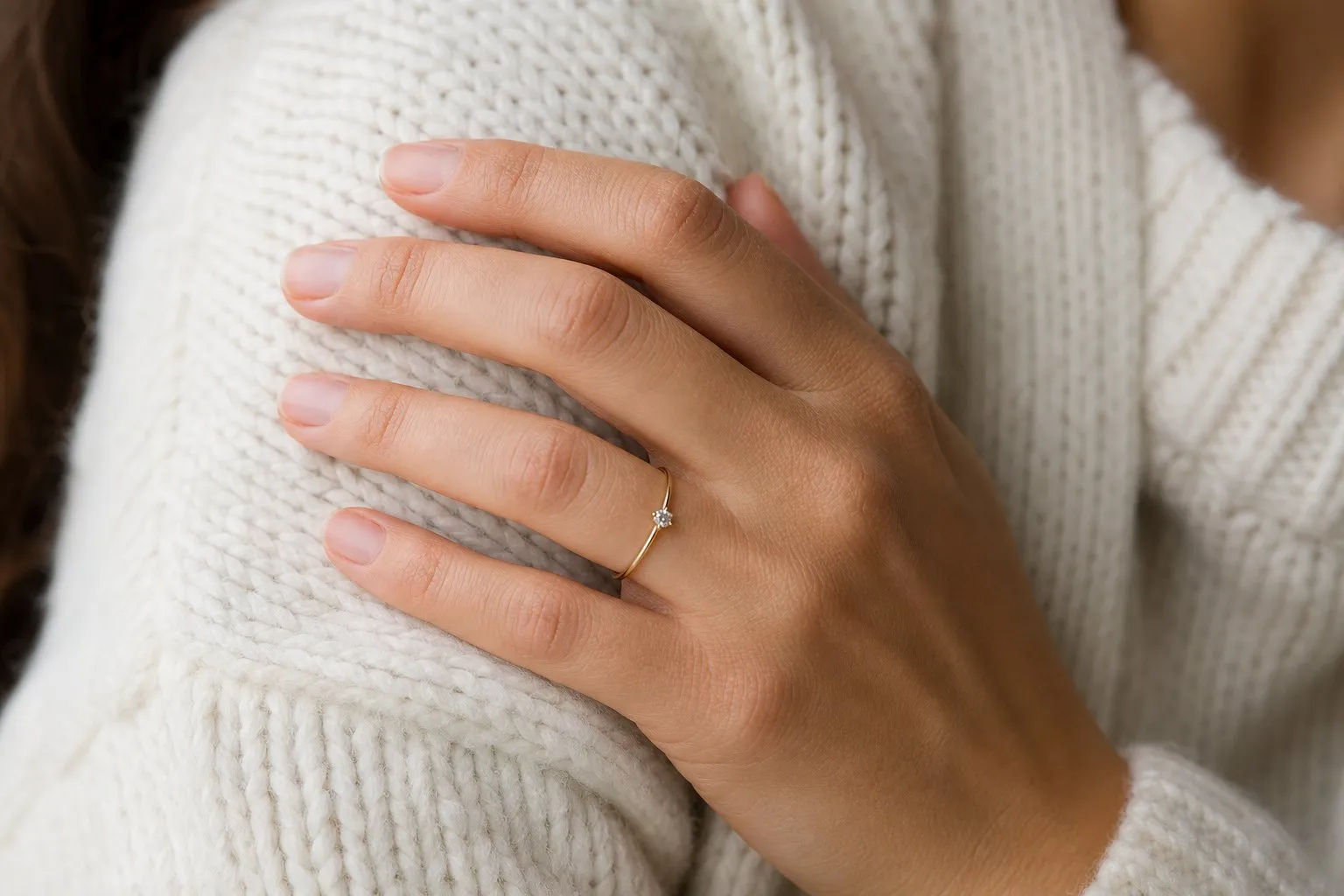 A hand resting on a cream knit sweater wearing a thin yellow gold solitaire engagement ring with a small round stone, the scale of stone found in complete engagement rings under $1,000