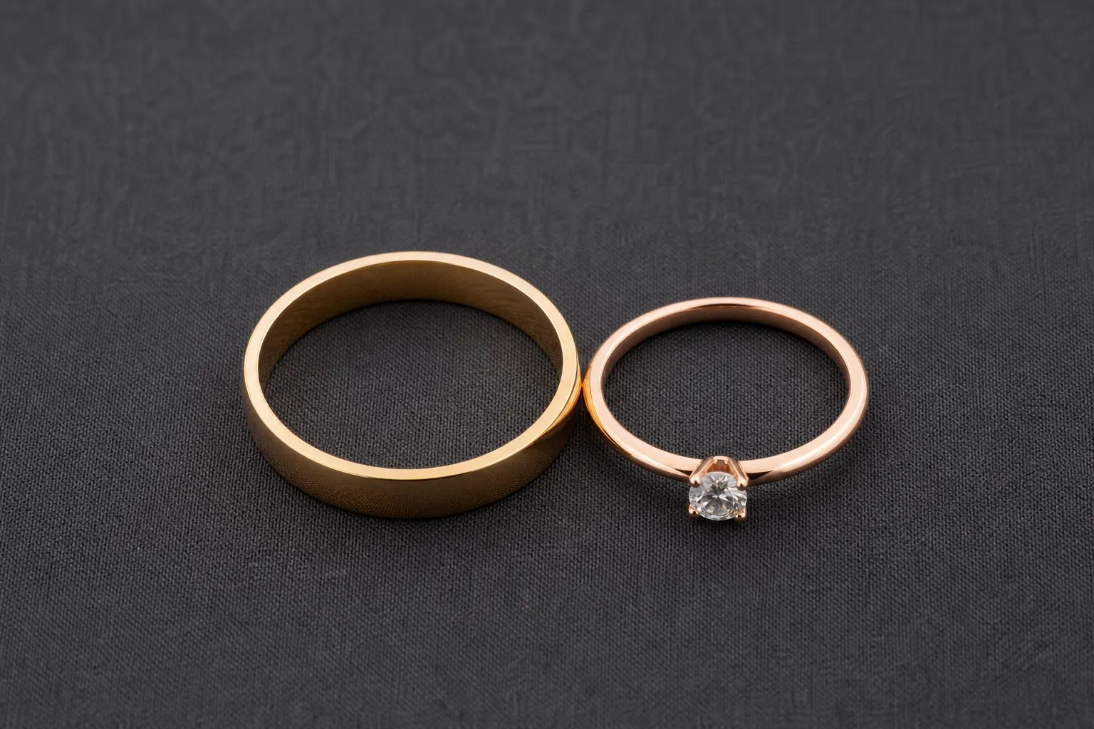 A flat band and a slim round-stone ring lying side by side on dark gray fabric, seen from above