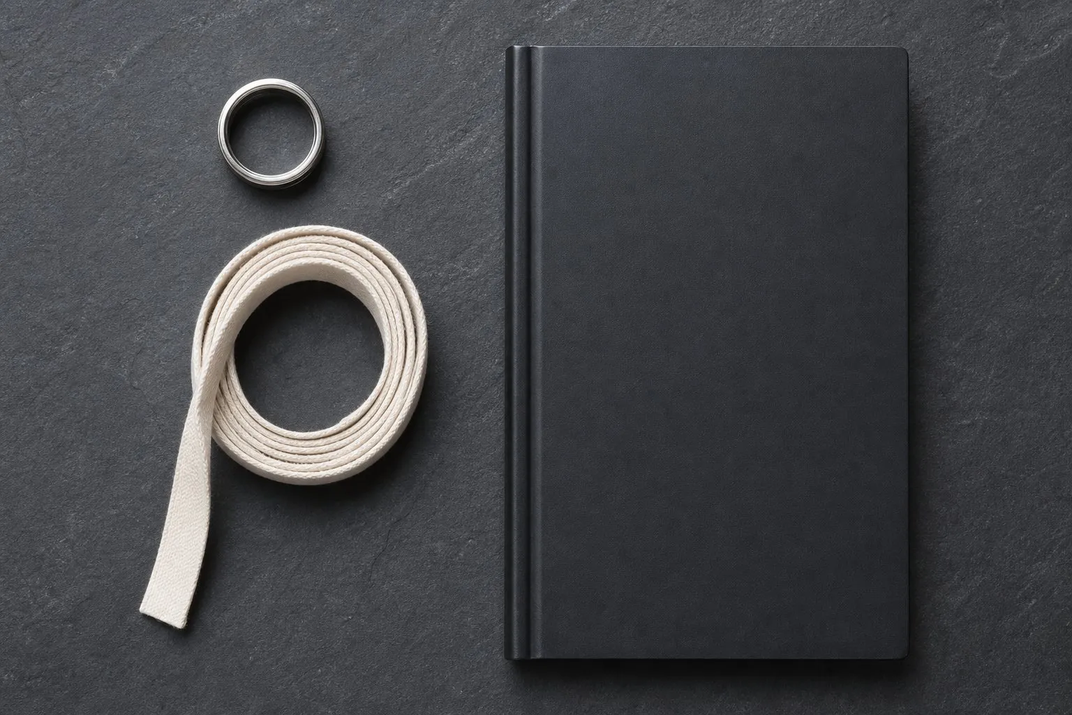 A plain metal ring, a coiled soft fabric tape and a closed black notebook on a slate gray surface