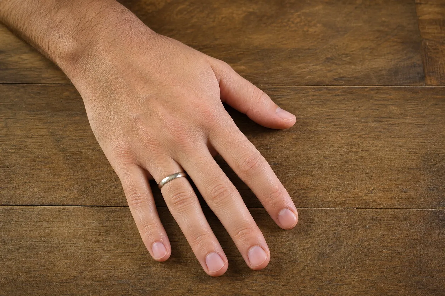 One hand lying flat on a wooden table wearing a single plain wedding band