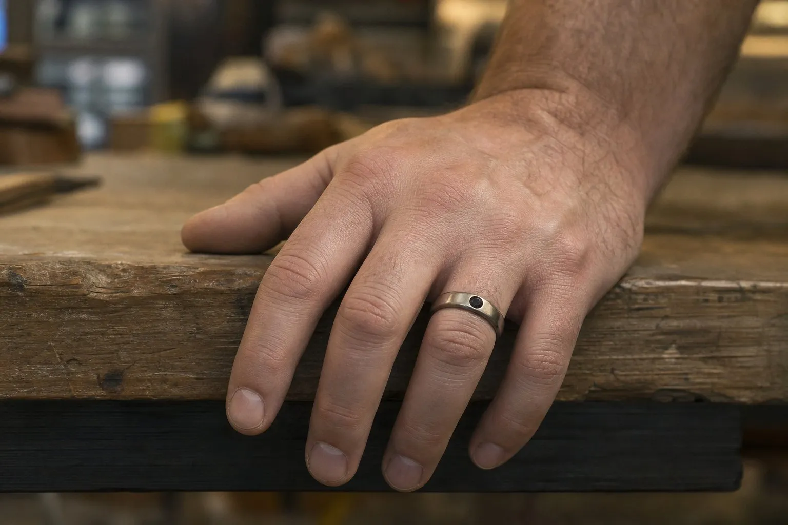 A man's hand gripping a workbench edge, wearing a low-profile band with a stone set level into the metal