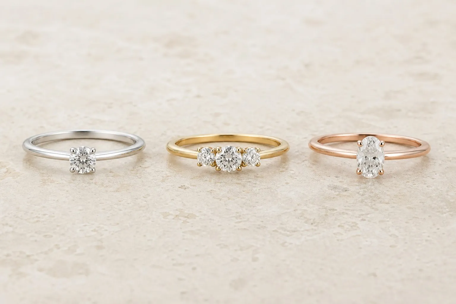 Three simple engagement rings in a row on an ivory stone surface: a white gold round solitaire, a yellow gold three-stone ring and a rose gold oval solitaire